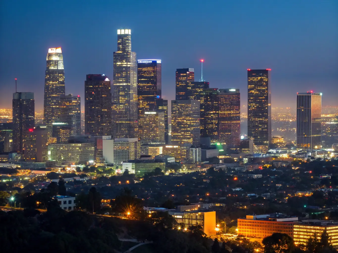 A nighttime aerial view of downtown Los Angeles, showcasing the city's skyline and bustling urban environment, with a subtle overlay of security camera icons to represent surveillance and protection.